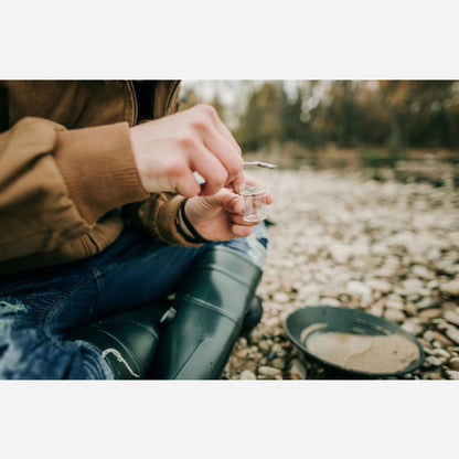 Person sitting outdoors by a stream, holding a small container.