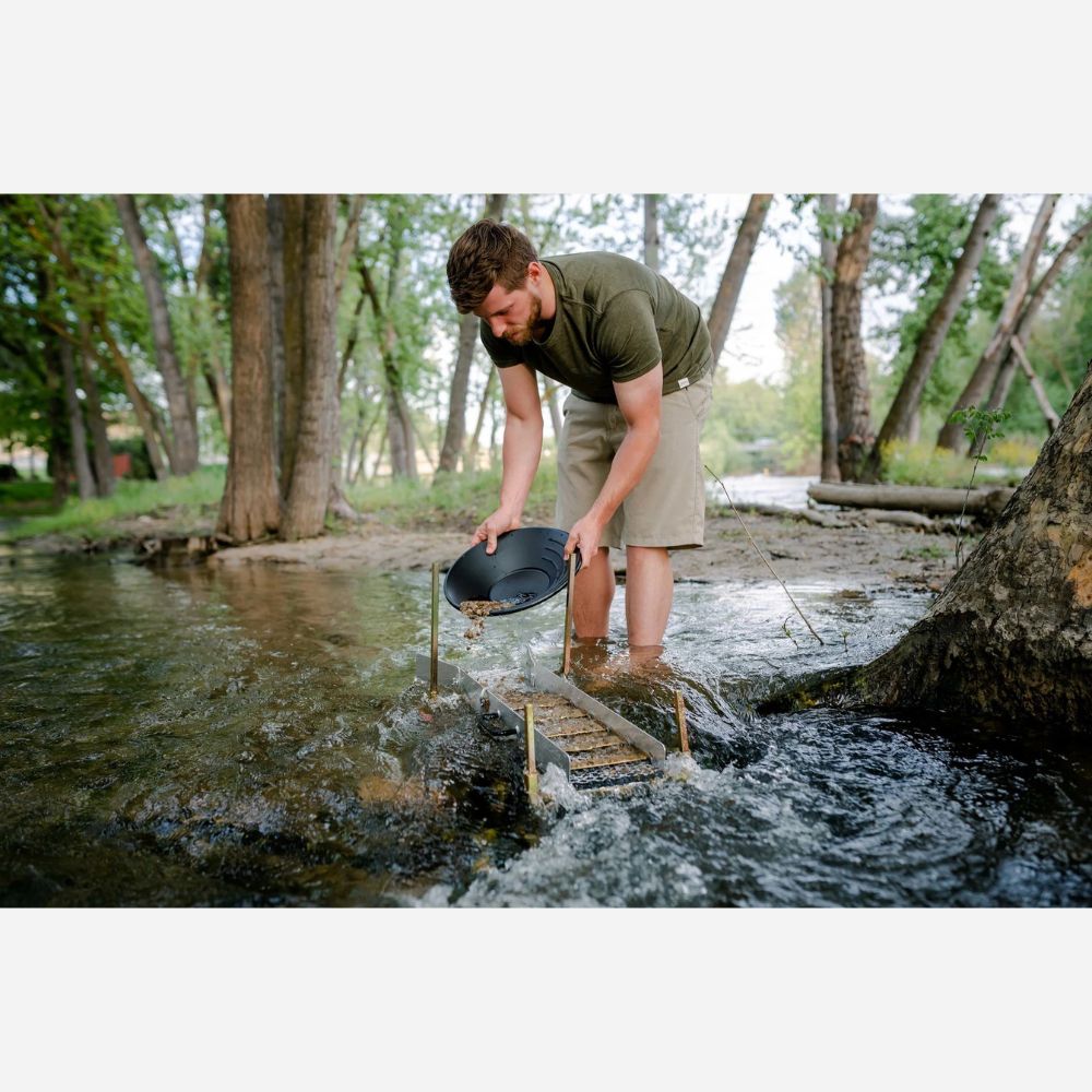 Man in a forest setting using a pan to sift through water, likely panning for gold.