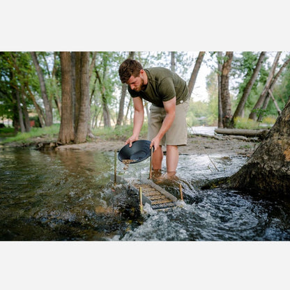 Man in a forest setting using a pan to sift through water, likely panning for gold.