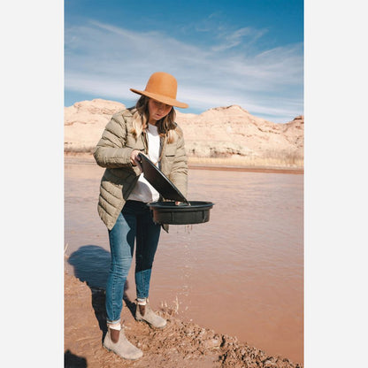Person standing by a lake with a hat and jacket, holding a metal pan.