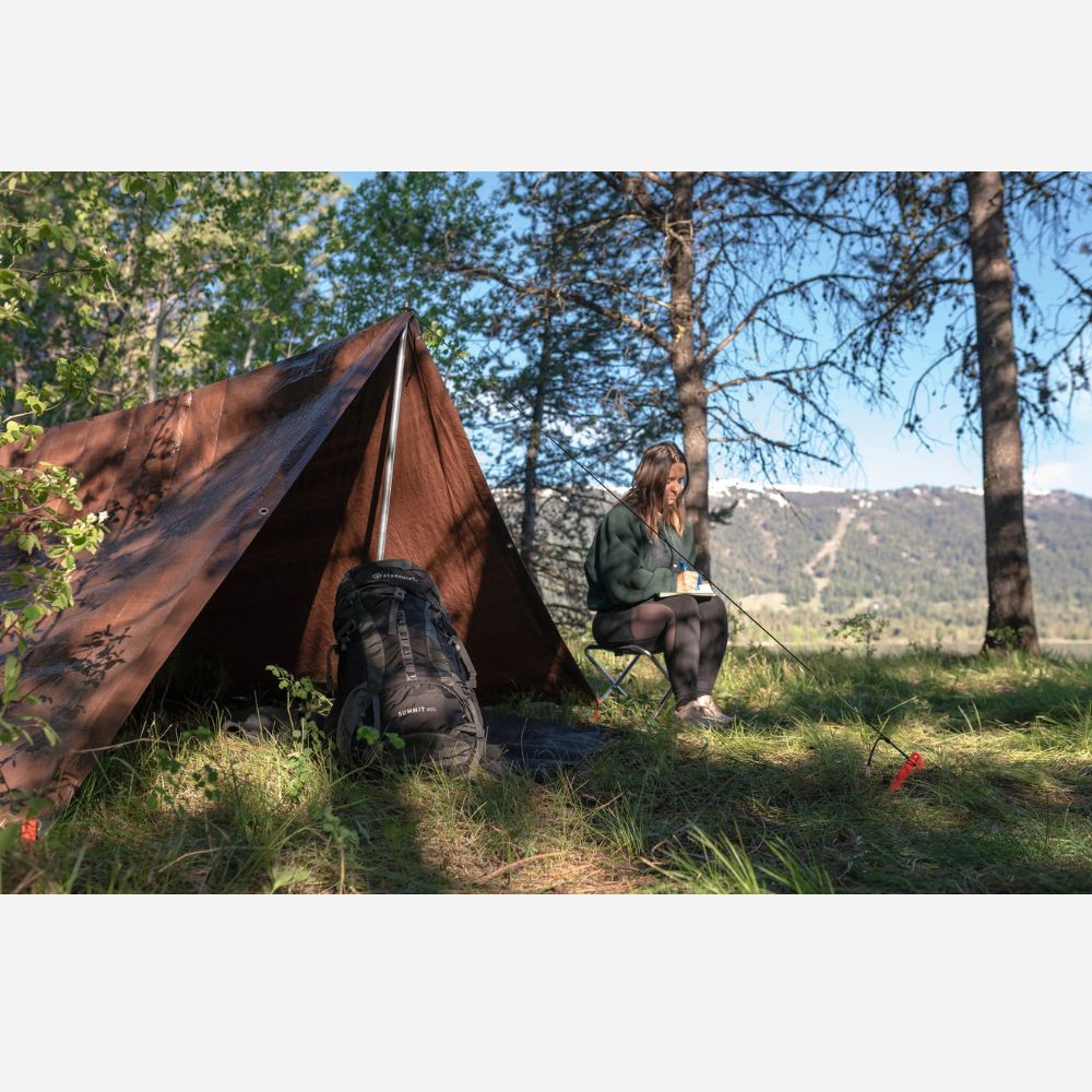 Person sitting by a tent in a forested area with mountains in the background