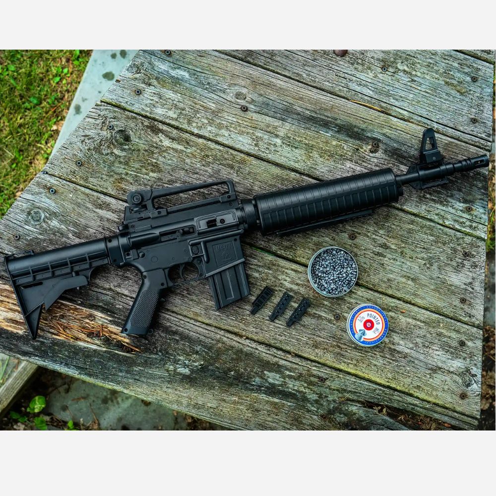 Black rifle on a wooden surface with two round objects and a circular object with a red dot.