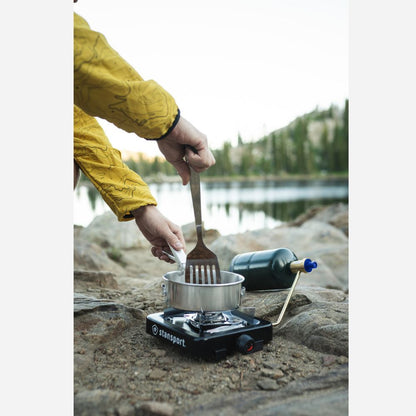 Person cooking outdoors using a portable stove by a lake