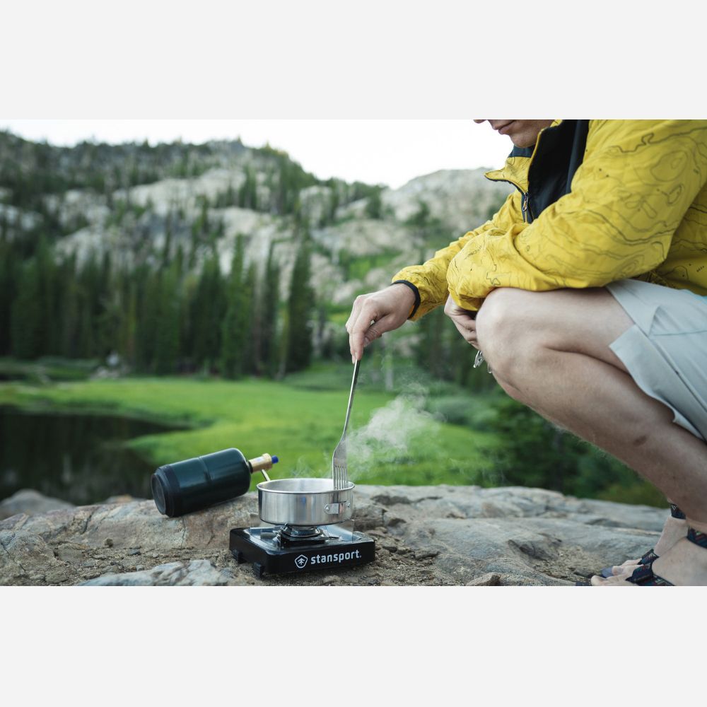 Person cooking outdoors using a camping stove with a scenic background