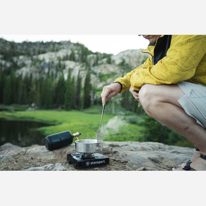 Person cooking outdoors using a camping stove with a scenic background