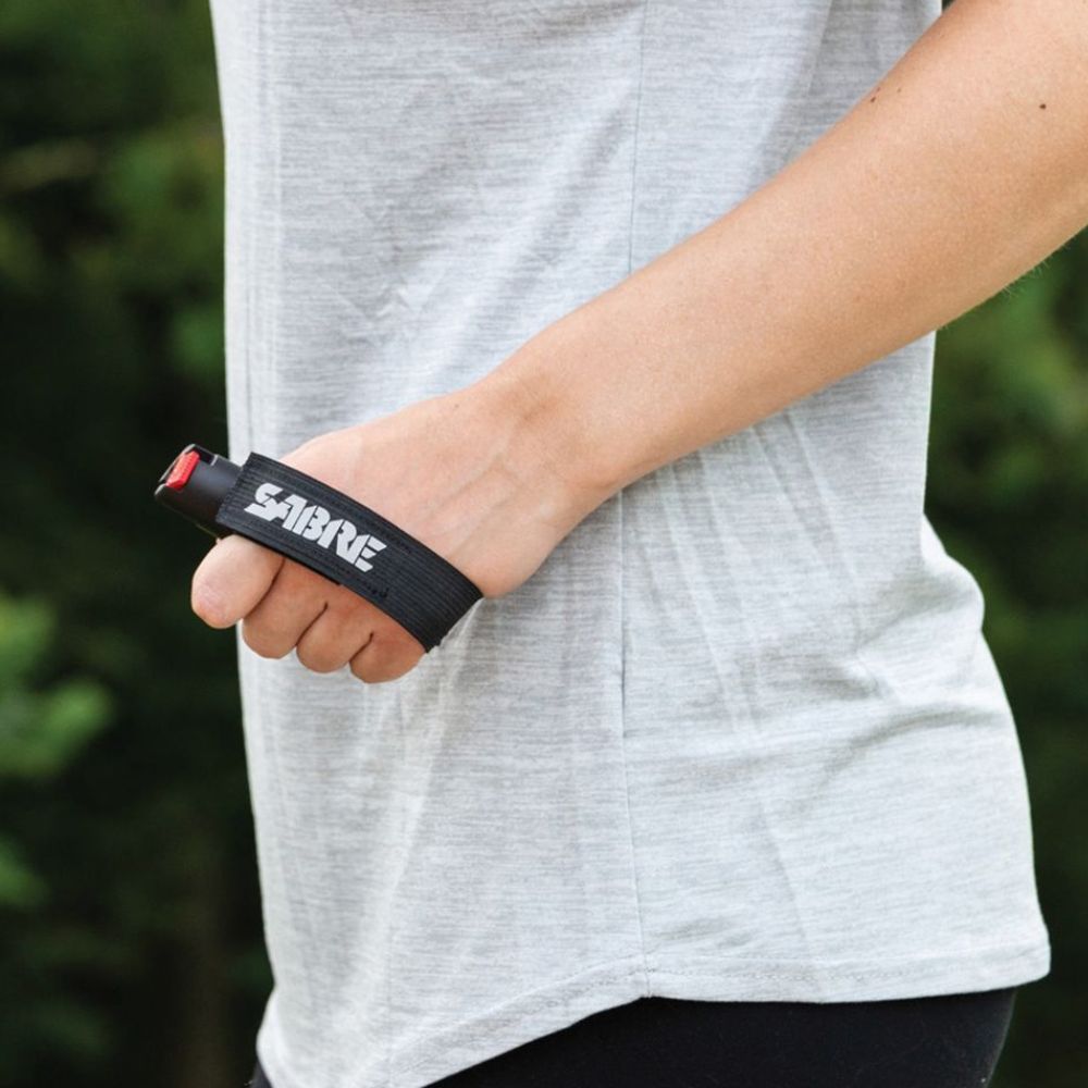 Person holding a black pepper spray device with 'Sabre' branding against a blurred green background