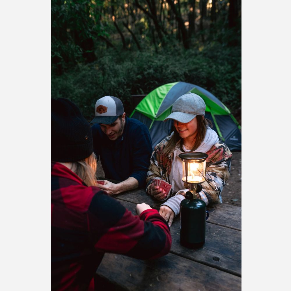 Three people camping outdoors, sitting around a lantern with a tent in the background.