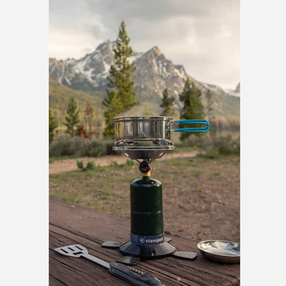 Camping stove with pot on a wooden surface against a mountainous landscape
