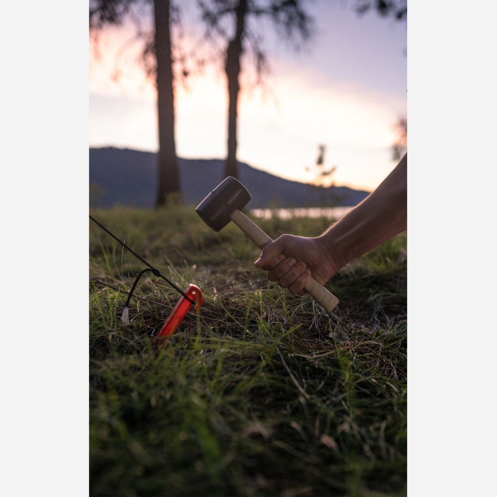 Hand holding a rubber mallet near a small red object on grass with trees in the background