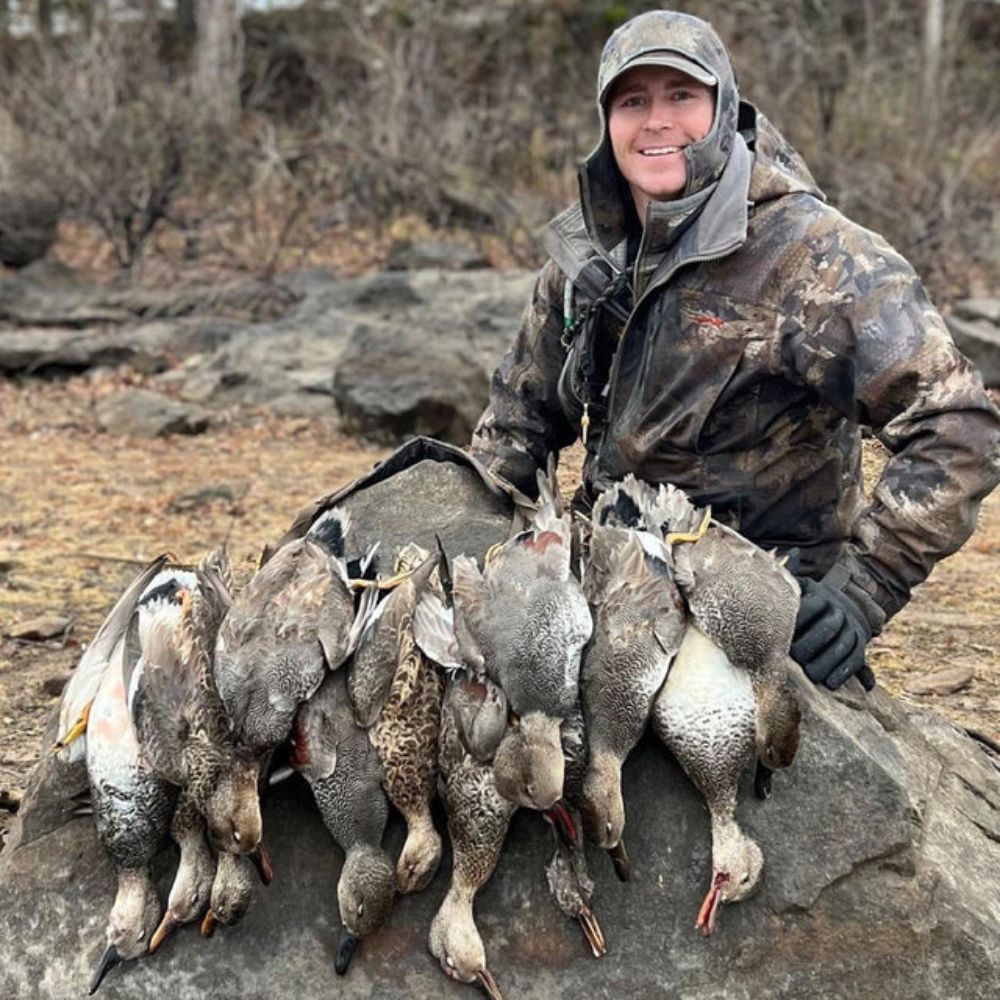 Person in camouflage clothing holding hunted birds on a rocky surface with a natural background