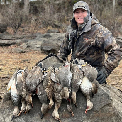 Person in camouflage clothing holding hunted birds on a rocky surface with a natural background