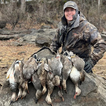 Person in hunting gear holding a pile of hunted ducks in a forest setting