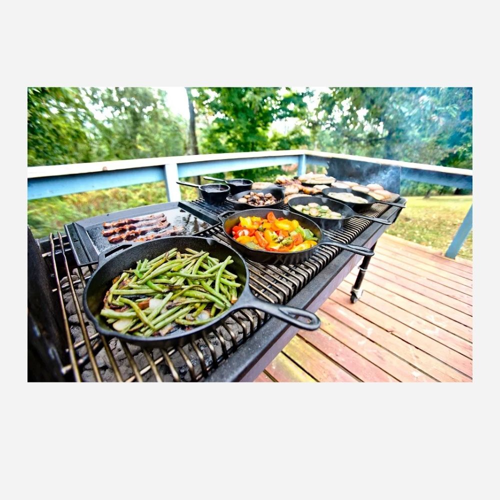 Outdoor barbecue grill on a wooden deck with cast iron skillets cooking green beans, mixed vegetables, mushrooms, and sliced meat over open flames.