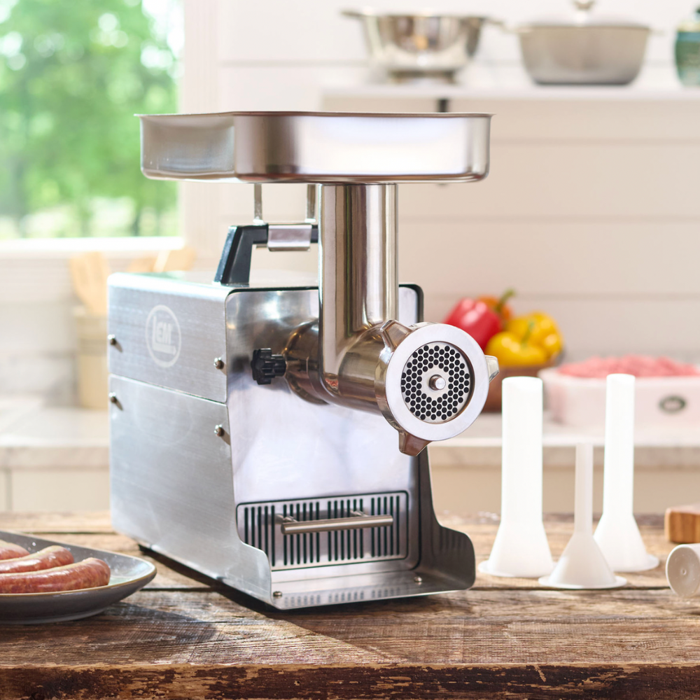 Stainless steel meat grinder on a kitchen counter with fruits and vegetables in the background