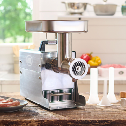 Stainless steel meat grinder on a kitchen counter with fruits and vegetables in the background
