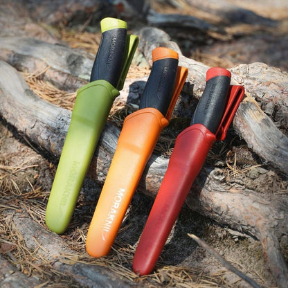 MoraKniv fixed blade knives in green, orange, and red sheaths displayed outdoors on tree roots