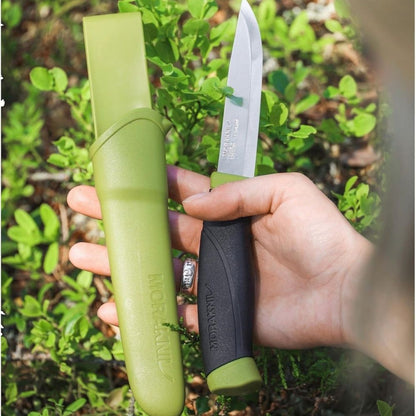MoraKniv fixed blade outdoor knife with green sheath and black handle held in hand outdoors among green foliage